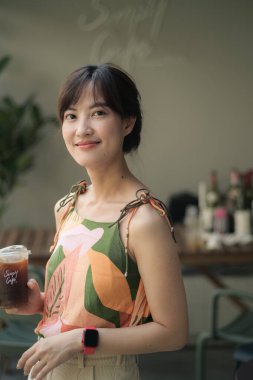Asyalı gülümseyen bir kadın açık hava kafe ortamında buzlu kahve tutuyor. Renkli kolsuz bluz ve şık elbiseler giyiyor.