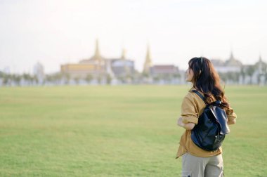 Sırt çantalı Asyalı bir kadın, arka planda bulanık tapınak yapıları olan çimen tarlasının huzurlu manzarasından zevk alıyor.