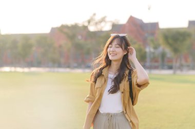 Neşeli bir Asyalı kadın açık havada güneşli bir günün tadını çıkarıyor. Rahat bir gülümsemeyle çimenli parkta duruyor. Günlük giysiler giyiyor ve sırt çantası taşıyor.