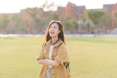 Neşeli Asyalı bir kadın, günlük kıyafetleriyle güneşli parkta kollarını kavuşturup, sıcak havanın keyfini çıkarıyor.