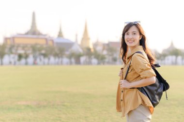 Sırt çantalı neşeli bir Asyalı kadın güneşli bir günün tadını çıkarıyor. Arka planda geleneksel mimari ve macera dolu bir hava var.