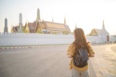 Sırt çantalı Asyalı bir kadın güneşli bir günde büyük tapınak kompleksinin önünde duruyor. Geleneksel mimariye ve sakin atmosfere hayran kalıyor.