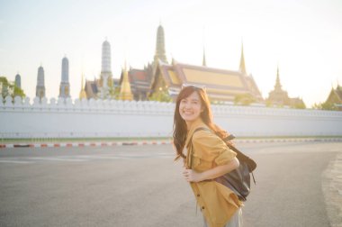 Sırt çantalı neşeli bir Asyalı kadın geleneksel tapınağın önünde güneşli bir günün tadını çıkarıyor. Altın kuleler, kültürel güzellikler ve seyahat titreşimleri sergiliyor.