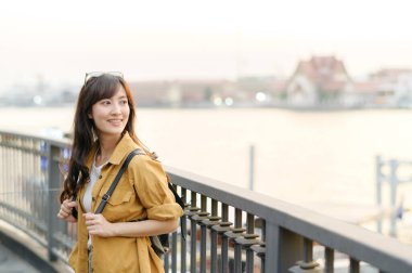 Sırt çantalı neşeli bir Asyalı kadın, gün batımında sakin nehir manzarasından zevk alır, hafif bir gülümsemeyle korkulukların yanında durur.