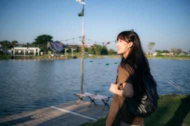 Sırt çantalı neşeli bir Asyalı kadın güneşli bir günün tadını çıkarıyor. Huzurlu bir göl, doğa ve huzurlu bir atmosfer ile çevrili.