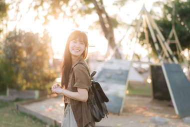 Sırt çantalı neşeli bir Asyalı kadın güneşli açık hava çocuk parkında gülümsüyor, yeşil ve sıcak güneş ışığıyla çevrili.
