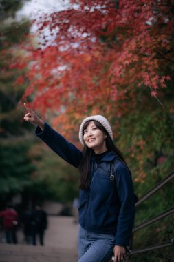 Neşeli Asyalı bir kadın rahat elbiseler giyerek yukarıyı gösterirken açık havada canlı sonbahar yeşilliklerinin tadını çıkarıyor.