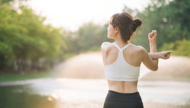Asyalı bir kadın kollarını açık havada, sakin park ortamında, atletik giysiler içinde, güneş ışığı ve yeşillik içinde, huzurlu bir atmosfer yaratıyor.