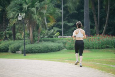 Parkta koşan Asyalı bir kadın. Etrafı yemyeşil, atletik kıyafetler giymiş, açık havada aktif ve sağlıklı bir yaşam sürüyor.
