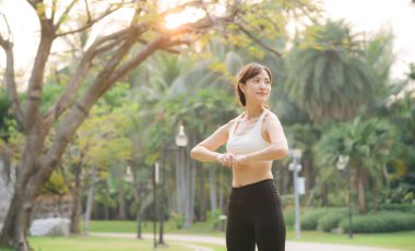 Parkta uzanan Asyalı bir kadın, beyaz spor bluz ve siyah tayt giyiyor, yemyeşil ve güneş ışığıyla çevrili.