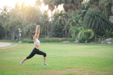 Huzur parkında yoga yapan Asyalı bir kadın, kollarını hareketli pozisyonda uzatıyor. Etrafı yemyeşil ve yumuşak güneş ışığı altında palmiye ağaçlarıyla çevrili.
