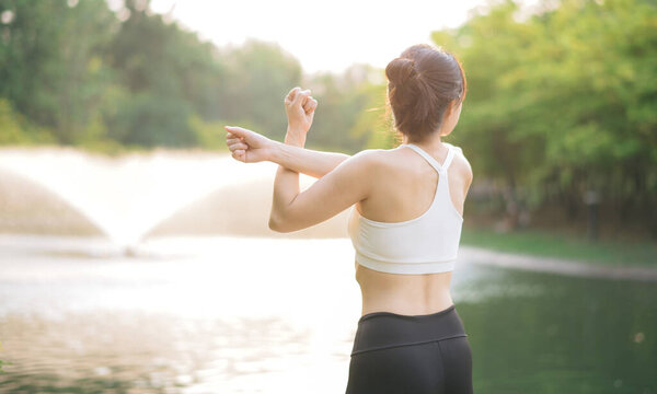 An Asian woman stretching her arms in serene park near pond, wearing white sports bra and black leggings, enjoying peaceful morning workout