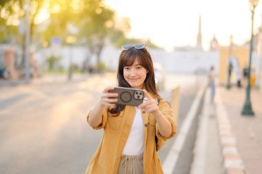 Gezgin Asyalı 30 'lu yaşlarda bir kadın Bangkok, Tayland' da şehir caddesinde dolaşırken fotoğraf çekmek için akıllı telefon kullanıyor.