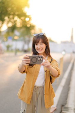 Gezgin Asyalı 30 'lu yaşlarda bir kadın Bangkok, Tayland' da şehir caddesinde dolaşırken fotoğraf çekmek için akıllı telefon kullanıyor.