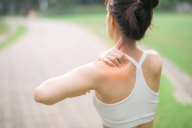 Omuz ağrısı sorunu. Kadın koşucu. 30 'lu yaşlarda beyaz spor giysili Asyalı bir kadın parkta koştuktan sonra omzunu acıyla tutuyor..