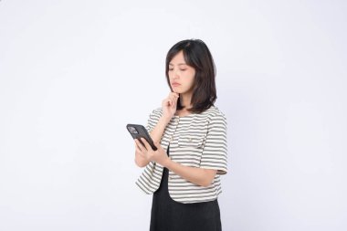 Güneydoğu Asyalı kadın internet uygulamalı cep telefonu kullanıyor. Düşünceli görünüyor ve sıradan çizgili ceket ve siyah elbiseyle beyaz arka plana odaklanıyor.
