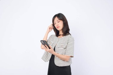 Güneydoğu Asyalı kadın internete düşünceli bir ifadeyle göz atmak için cep telefonu kullanırken, günlük kıyafetlerin içinde sade beyaz arka plana karşı duruyor.