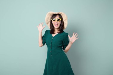 Sarı güneş gözlüğü ve hasır şapka takan neşeli güneydoğu Asyalı kadın yeşil elbiseyle arka planda reklam yapmak için mutlu bir şekilde poz veriyor.