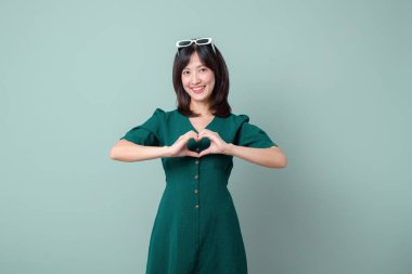 Yeşil elbiseli Güneydoğu Asyalı kadın gülümsüyor ve elleriyle kalp şekli oluşturuyor. Neşeli bir ifadeyle reklamını yapıyor.