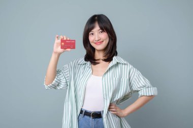 Çevrimiçi alışveriş yaparken güvenle gülümseyen Güneydoğu Asyalı kadın kredi kartını elinde tutuyor. Günlük çizgili gömlek ve kot pantolon ile ödemeye veya satın almaya hazır.