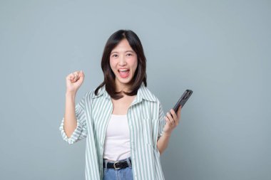 Mutlu Güneydoğu Asyalı kadın internet bağlantısı olan cep telefonu kullanıyor. Sade arka planda neşeli ifadesi olan günlük kıyafetlerle mobil uygulamanın tadını çıkarıyor.