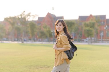 Sırt çantalı Güneydoğu Asyalı kadın güneşli park ortamında gezmekten keyif alıyor. Neşeli bir ifade takınıyor. Günlük kıyafet ve doğal ışık ile seyahat kavramını yakalıyor.