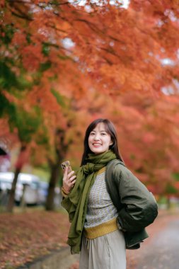 Güneydoğu Asyalı kadın, arka planda canlı turuncu yapraklarla sonbaharda açık havada telefonla konuşurken sıcak ve neşeli bir atmosfer yaratarak gülümsüyor.