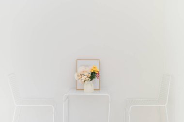 Çiçek aranjmanlı küçük bir masa, çerçeveli bir resim ve temiz ve sakin bir atmosfer yaratan iki beyaz sandalye içeren minimalist beyaz bir oda.