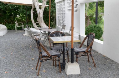 Beyaz duvarlar ve yeşil bitkilerle çevrili, büyük çakıllı zeminde siyah ve beyaz dokuma sandalyeleri ve küçük şemsiyeli küçük masaları olan bir açık hava oturma alanı barışçıl atmosfer yaratıyor.