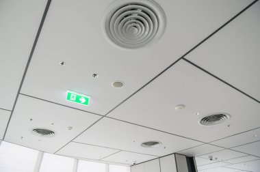Temiz, modern ofis tavanında hava delikleri, aydınlatma ve ızgara deseni, yeşil parlayan acil çıkış işareti, parlak ve düzenli bir atmosfer yaratıyor.