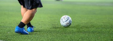 Mavi kramponlu bir futbolcu yeşil çimlerde beyaz topla, güneşli bir günde canlı renkler ve net detaylarla hareket ve odaklanma gösterir.