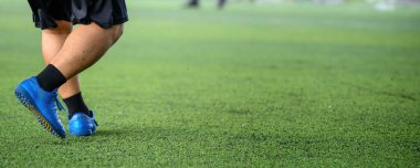 Maç sırasında yeşil çimlerde mavi krampon ve siyah çorap giyen bir futbolcu, canlı renkler ve keskin detaylarla hareket halinde olan alt vücut ve ayakkabılara odaklanıyor