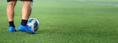 Yeşil çimlerin üzerinde mavi kramponlar ve siyah çoraplarla duran bir futbolcu, maç sırasında ya da antrenman sırasında ayağa yakın topa odaklanıyor