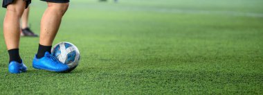 Maç sırasında yeşil çimlerde topu kontrol eden mavi krampon takan bir futbolcu, açık hava spor ortamında bacaklara ve top hareketlerine odaklanıyor