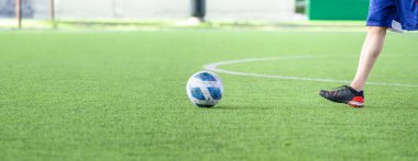 Siyah krampon ve mavi şort giymiş, güneşli bir günde topa vurmaya hazırlanan bir futbolcuyla yeşil çimlerde bir futbol topu.