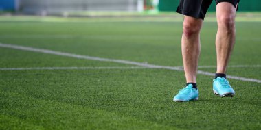 Yeşil çimlerin üzerinde duran bir futbolcu mavi kramponlar ve siyah şortlar giyerek ayaklara ve ayakkabılara odaklanmış spor oyunlarında harekete geçmeye hazır.
