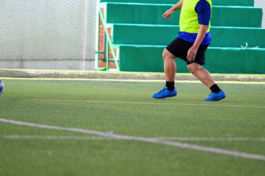 Mavi ayakkabılar ve siyah şortlar giyen bir futbolcu oyun sırasında yeşil suni çim sahasında koşuyor ve top yakınlarda, spor hareketlerini ve hareketlerini gösteriyor.