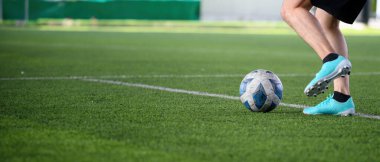 Mavi krampon takan bir futbolcu maç sırasında yeşil çimlerde beyaz ve mavi topa vurmak üzere, bacaklarına ve topa odaklanıyor.