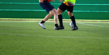 Futbol oyuncuları çim sahası boyunca hareket halindedir. Ayaklarına ve toplarına odaklanırlar. Dinamik hareket ve rekabet yoğunluğunu gösterirler.