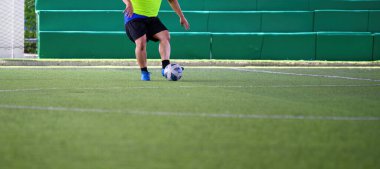 Maç sırasında yeşil çimlerde topu kontrol eden bir futbolcu, açık sarı gömlek ve siyah şort giyiyor, mavi kramponlarla harekete odaklanıyor.