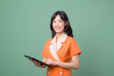 Güneydoğu Asyalı bir kadın internet tableti kullanıyor turuncu elbiseli gülümsüyor yeşil arka plan modern teknoloji konseptine karşı duruyor