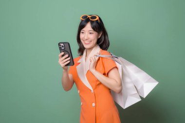 Güneydoğu Asyalı bir kadın, elinde alışveriş çantaları ve akıllı telefonuyla yeşil arka plandaki neşeli ifadeye gülümsüyor.