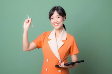 Turuncu üniformalı, elinde dijital tablet ve stil kalemle gülümseyen güneydoğulu bir kadın yeşil arka planda durarak güven ve pozitiflik gösteriyor.