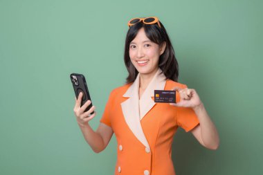 Turuncu elbiseli güneydoğulu bir kadın elinde akıllı telefon ve kredi kartıyla yeşil arka plana gülümsüyor. Modern yaşam tarzı.