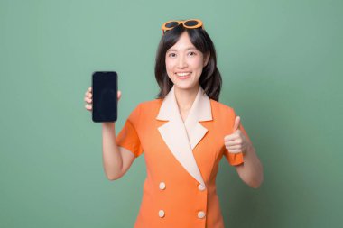Turuncu elbiseli güneydoğu Asyalı bir kadın elinde akıllı telefon ve yeşil arka planda mutlu bir ifadeyle başparmağını kaldırıyor.