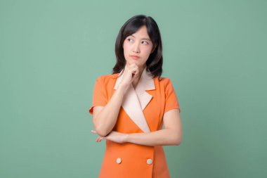 Turuncu elbiseli güneydoğu Asyalı bir kadın yeşil arka plana karşı düşünceli bir ifadeyle duruyor, elleri çenesinde, yukarı bakıyor, merak ve düşünce gösteriyor.