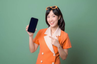 Güneydoğu Asyalı bir kadın, turuncu elbiseli ve güneş gözlüklü, elinde akıllı telefonla gülümseyen ve yeşil arka planda boş ekran olan bir kadını işaret ediyor.