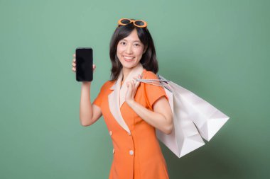 Güneydoğu Asyalı, turuncu elbiseli, elinde akıllı telefon ve alışveriş torbalarıyla gülümseyen bir kadın yeşil arka plandaki neşeli ifadeye karşı.