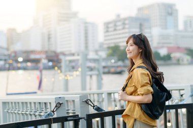 Güneydoğu Asyalı uzun saçlı, günlük sarı tişörtlü ve sırt çantalı bir kadın şehrin nehir kenarında durup gülümsüyor ve şehir dışı seyahatlerinin tadını çıkarıyor.