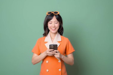 Güneydoğu Asyalı, turuncu elbiseli, gülümseyen bir kadın. İnterneti yeşil arka plana karşı kullanırken mutluluğu ve modern yaşam tarzını ifade ediyor.